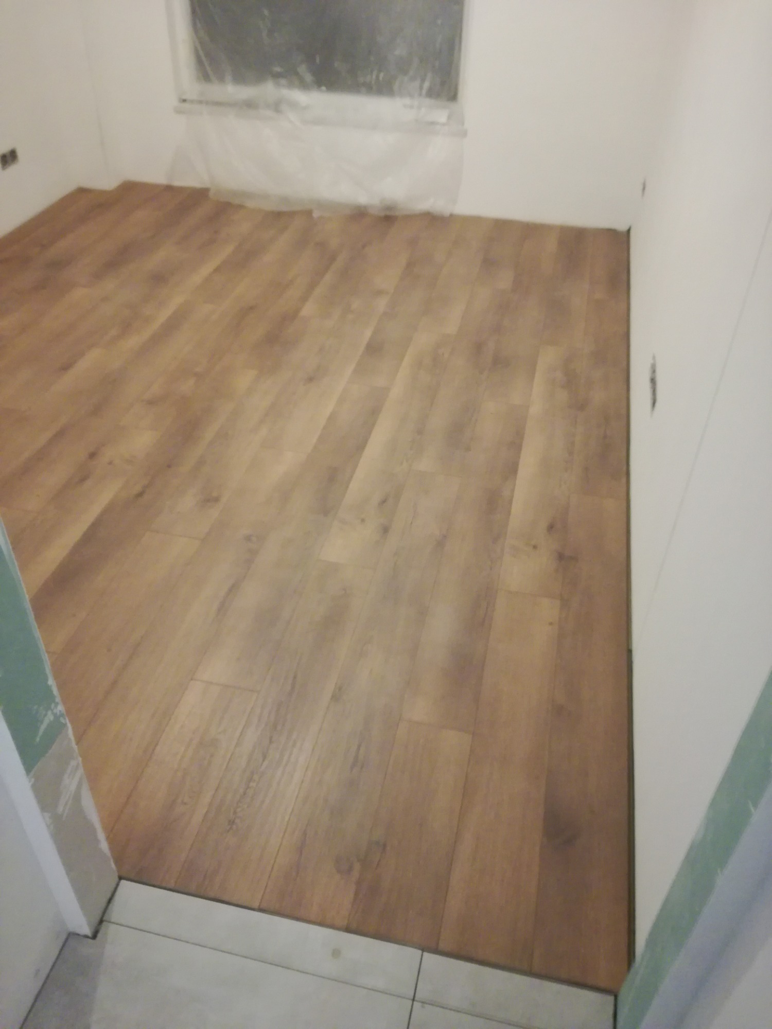 Ułożone panele podłogowe w pokoju, widoczne łączenia, przy ścianie biała listwa, okno zabezpieczone folią malarską.