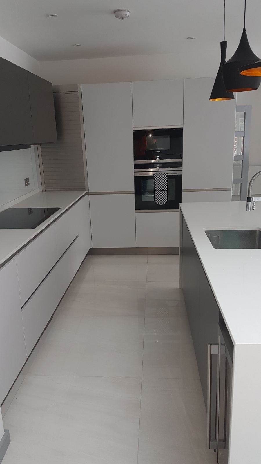 Nowoczesna kuchnia w Legionowie z białymi i szarymi szafkami, czarną płytą grzewczą i piekarnikiem, minimalistycznym oświetleniem oraz jasną podłogą z płytek.
