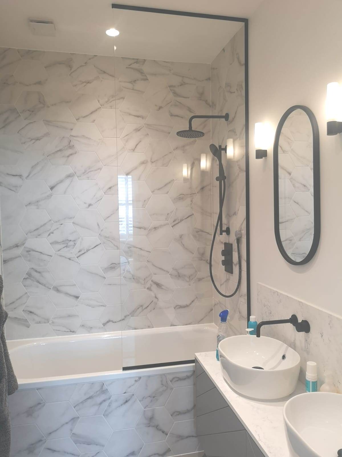 Wykończona łazienka z biało-szarymi heksagonalnymi płytkami imitującymi marmur, czarną armaturą i owalnym lustrem, z widoczną wanną i umywalkami nablatowymi.