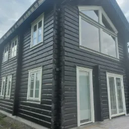 Ciemna, drewniana elewacja domu z jasnymi ramami okiennymi i dużym oknem trójkątnym na poddaszu, widok z dołu.