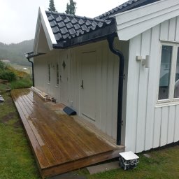 konrad jankowski - Biały, drewniany budynek z czarnym dachem po deszczu; widoczny mokry drewniany taras, puszki z farbą i narzędzia malarskie sugerujące prace wykończeniowe.