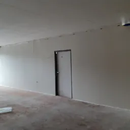 Wnętrze w trakcie remontu: gładkie, jasne ściany, surowy beton na podłodze, widoczne przewody elektryczne pod sufitem i fragment drabiny malarskiej w rogu.