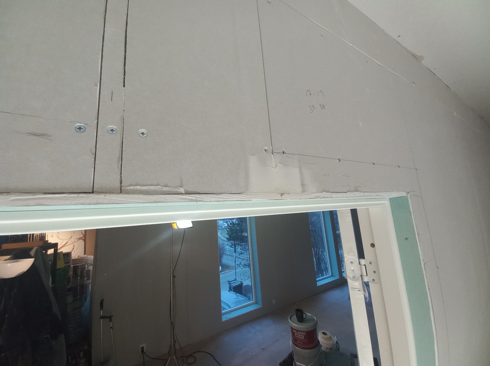 Montaż płyt gipsowo-kartonowych na ścianie nad ościeżnicą drzwiową, widoczne ślady ołówka, wkręty, szczeliny między płytami i wiadro z masą szpachlową w tle.