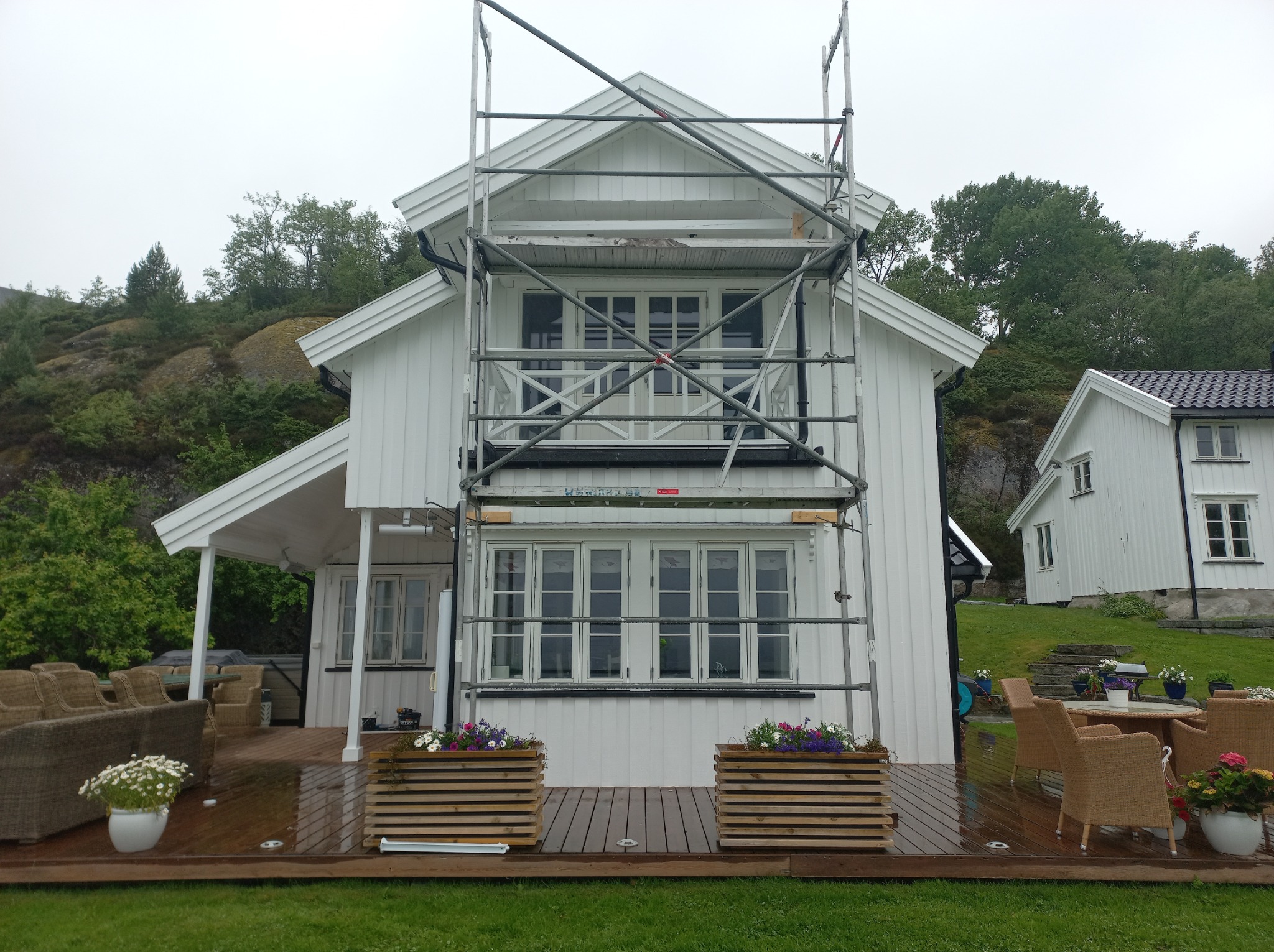 Biały dom z rusztowaniem podczas malowania elewacji, widoczne świeżo pomalowane fragmenty ściany i drewniane donice z kwiatami na tarasie.