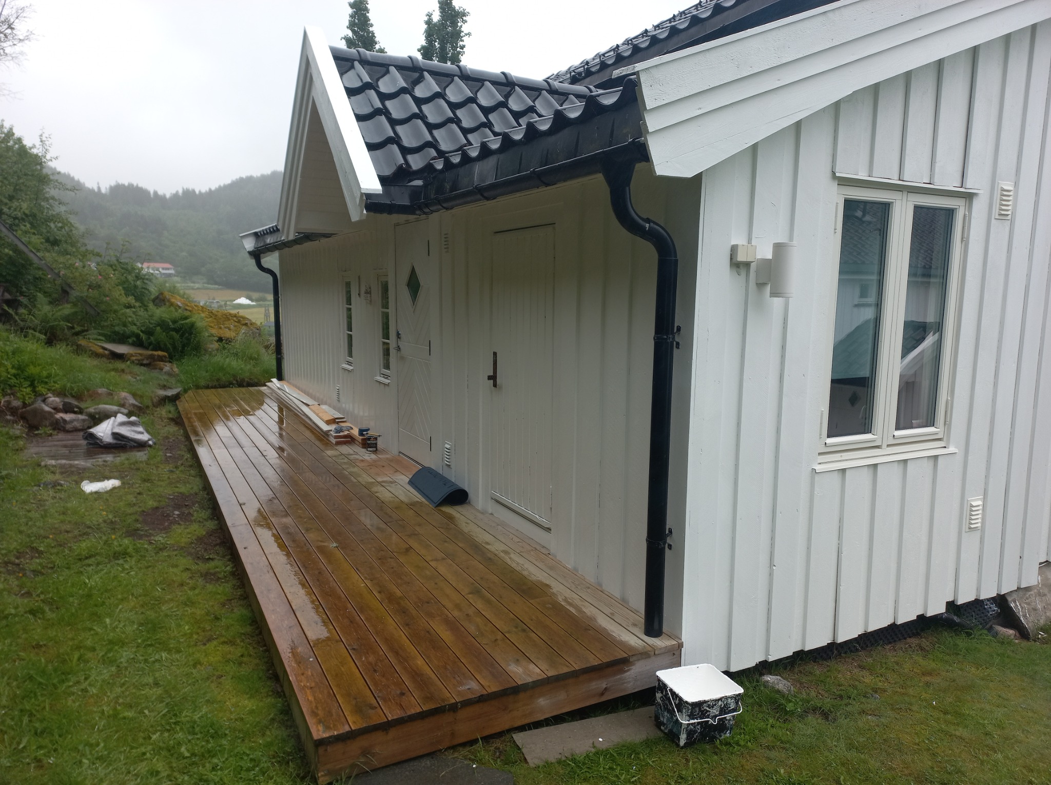 Biały, drewniany budynek z czarnym dachem po deszczu; widoczny mokry drewniany taras, puszki z farbą i narzędzia malarskie sugerujące prace wykończeniowe.