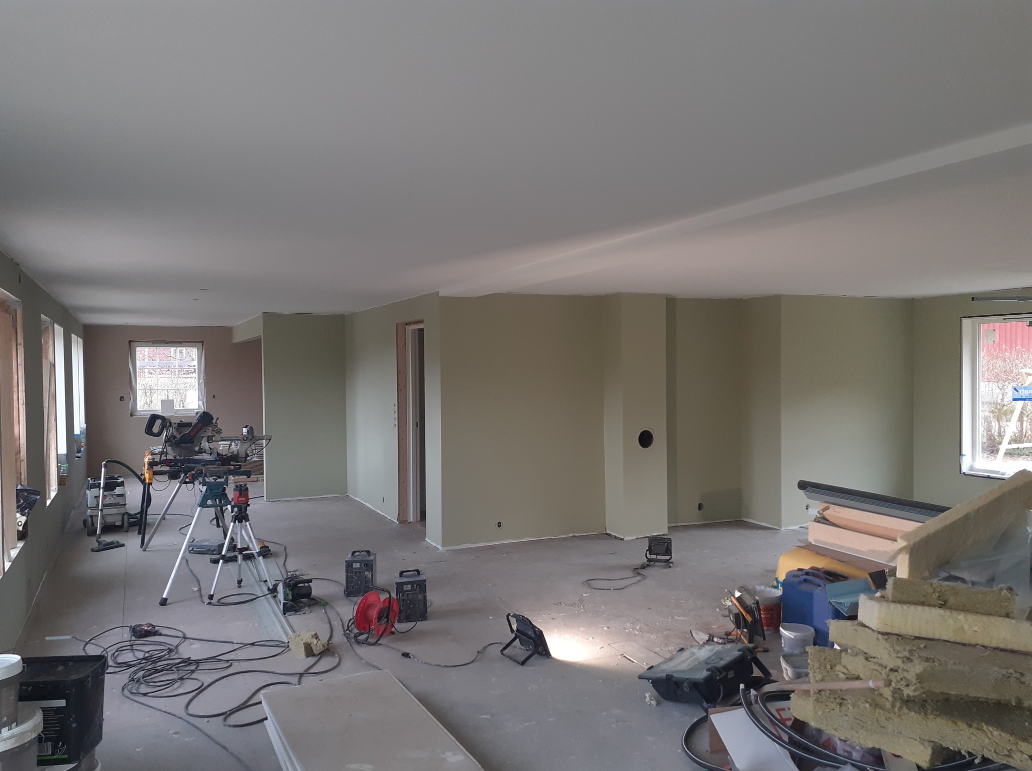 Duże, jasne wnętrze w trakcie prac wykończeniowych; ściany pomalowane na oliwkowy kolor, widoczne narzędzia, kable, lampy budowlane, resztki materiałów budowlanych, w tle okna.