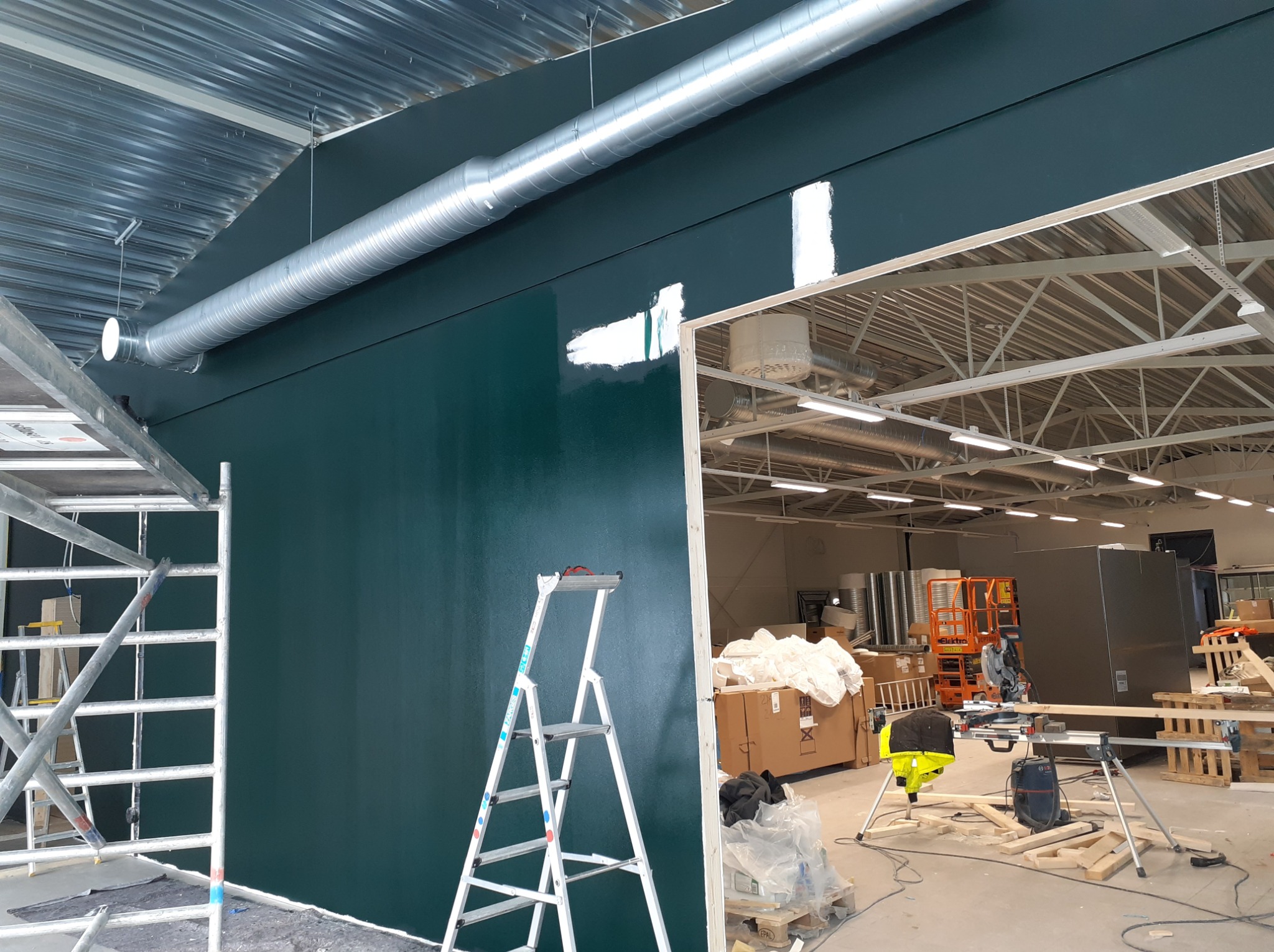 Świeżo pomalowana na ciemnozielono ściana w dużym pomieszczeniu przemysłowym, z widocznymi śladami po szpachlowaniu i częściowo pomalowanymi fragmentami, drabiny malarskie i rusztowanie w pobliżu.