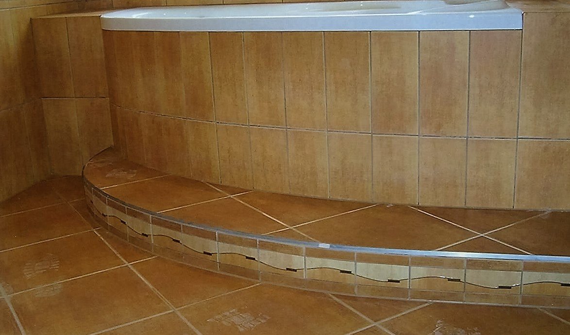 Wykończenie wnęki wanny z zaokrąglonym stopniem, wyłożone brązowymi płytkami ceramicznymi z dekoracyjnym pasem mozaiki.