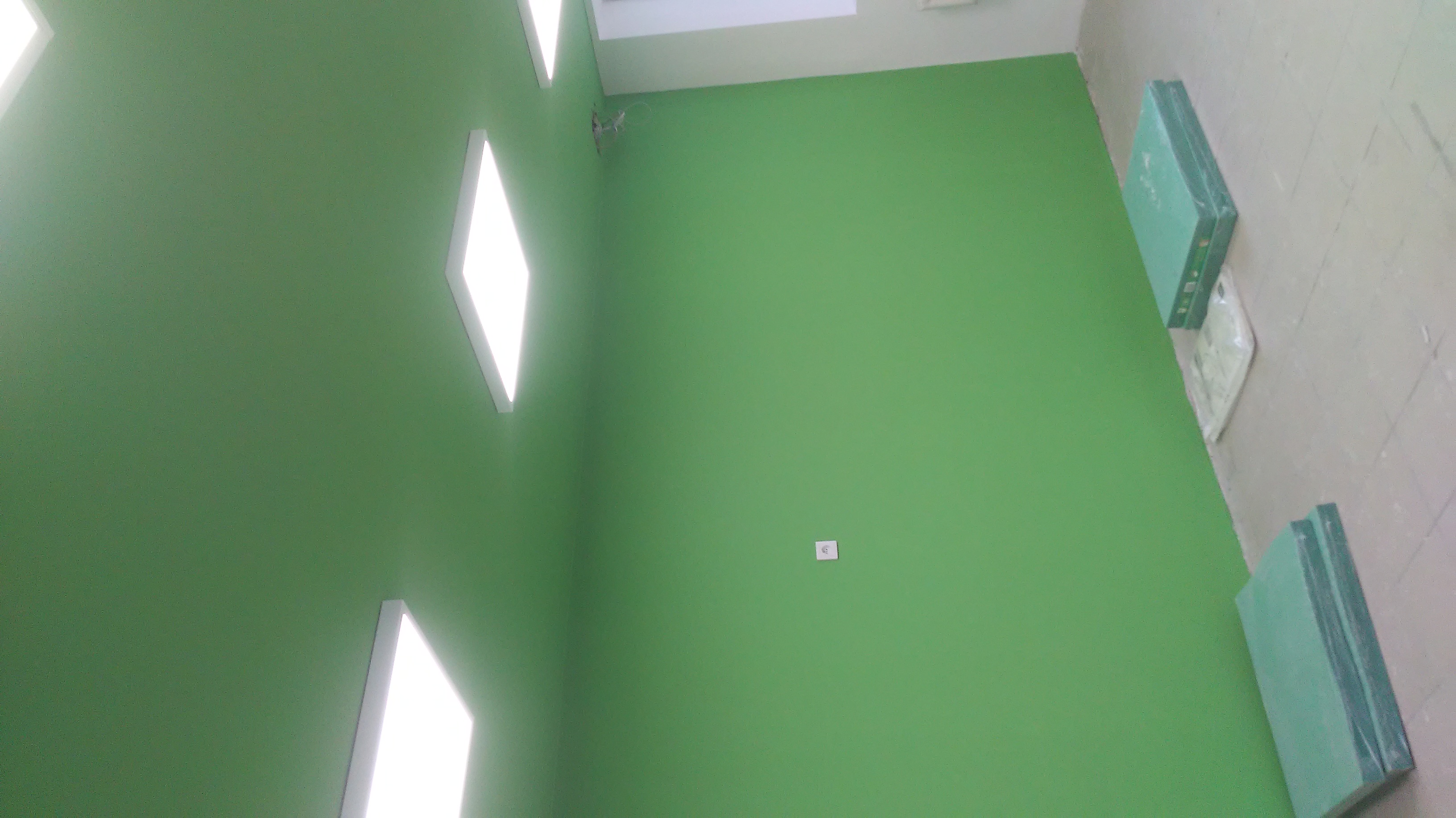 Świeżo pomalowana, jednolita, jasnozielona ściana w biurze w Rudzie Śląskiej, oświetlona przez trzy podłużne lampy LED. Na podłodze, przy ścianie, leżą paczki płyt gipsowo-kartonowych.