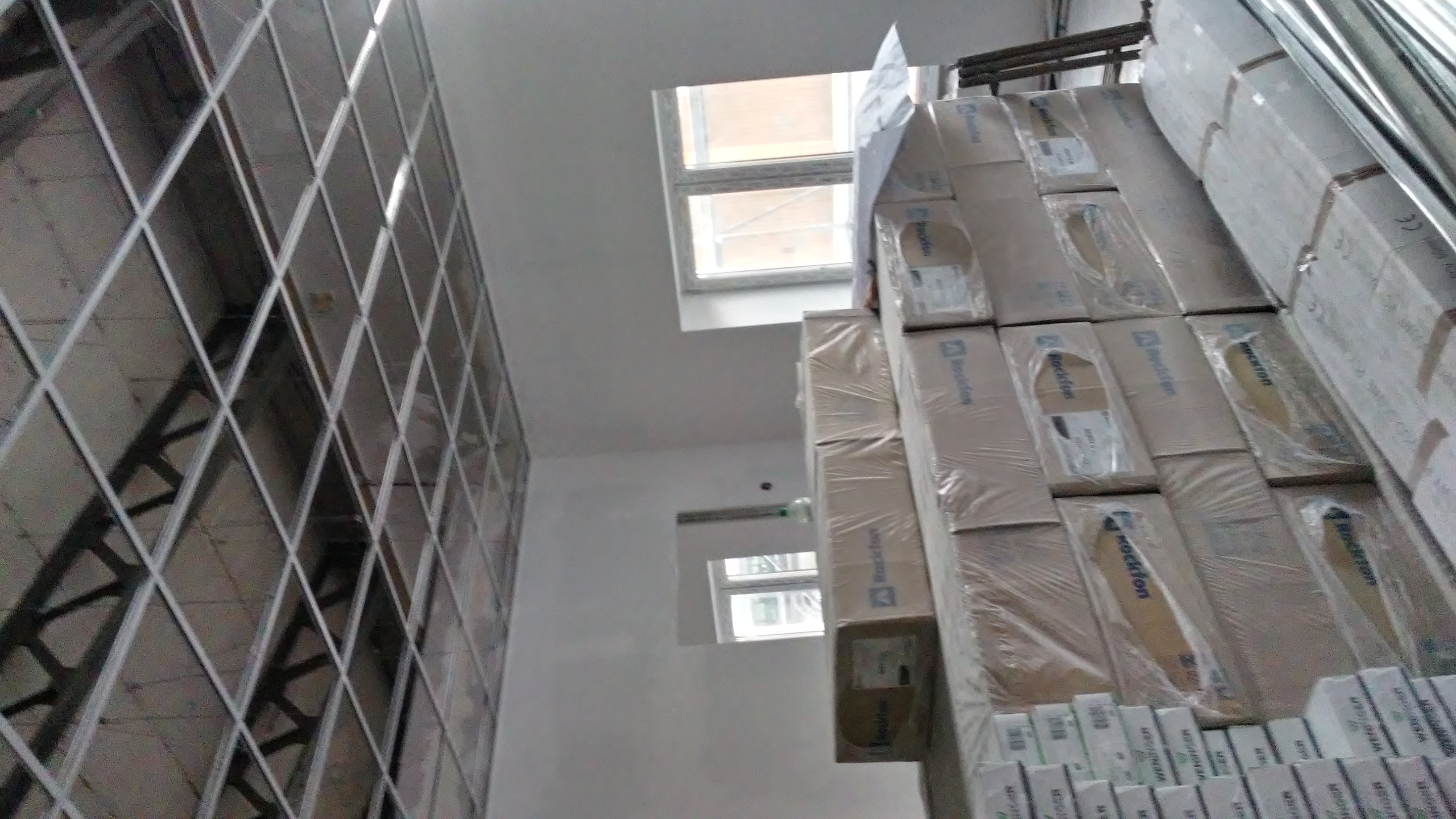 Metalowa konstrukcja szkieletowa ściany z płyt kartonowo-gipsowych, widok od dołu, obok stos zapakowanych płyt Rockfon, okna w tle.