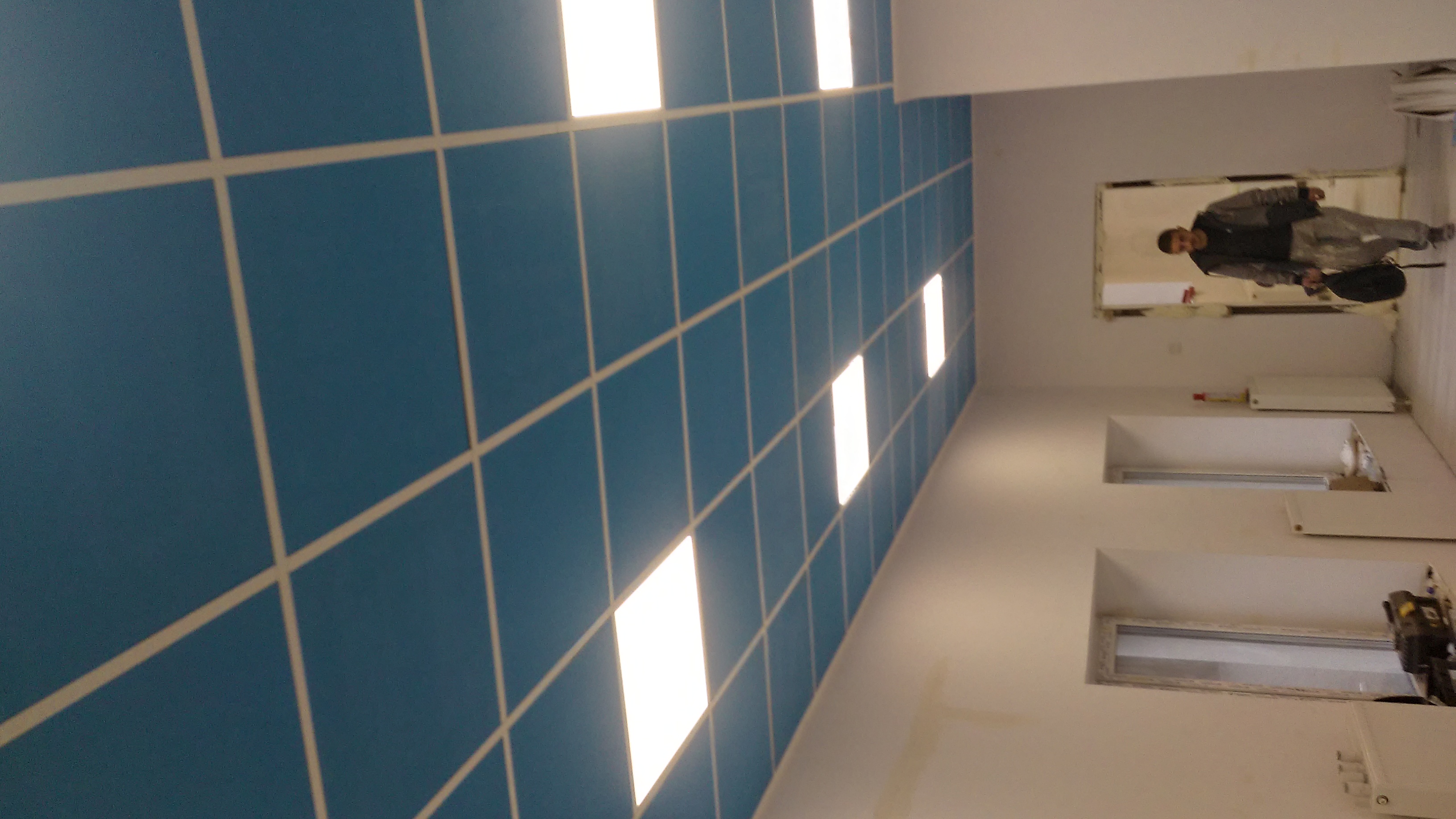 Niebieski sufit kasetonowy z białymi liniami podziału i wbudowanym oświetleniem w remontowanym biurze, widoczny pracownik w tle.