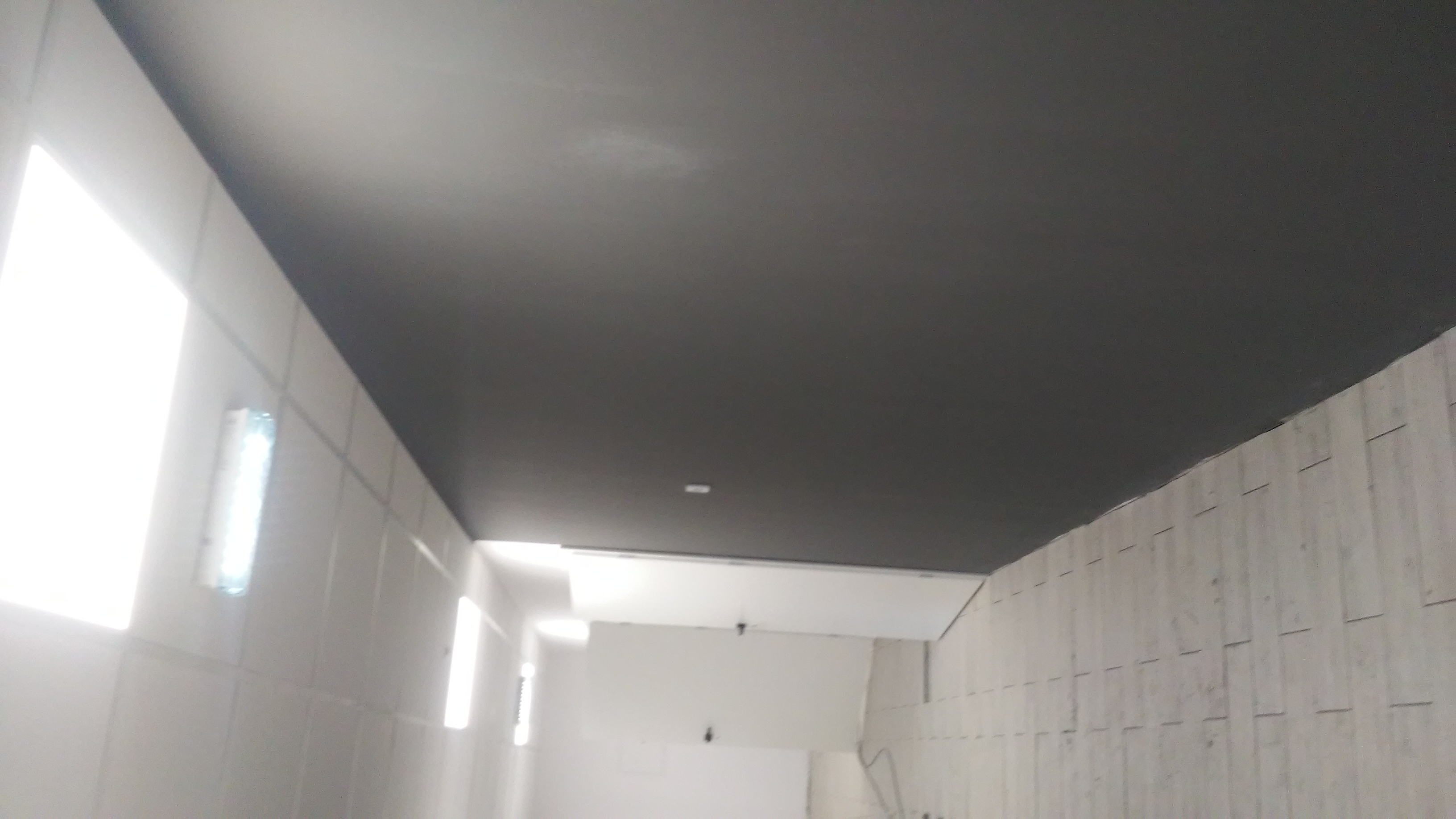 Pomieszczenie po malowaniu sufitu na ciemny szary kolor, ściany w jasnych odcieniach, widoczne oświetlenie jarzeniowe.