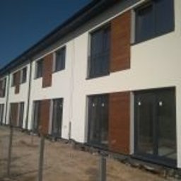 ARBUD Artur Buza - Elewacja szeregowca z zamontowanymi oknami PCV i drewnianymi panelami. Jasna fasada, ciemne okna, minimalistyczny design. Widok z zewnątrz, w tle błękitne niebo.