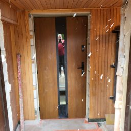 ARBUD Artur Buza - Montaż brązowych drzwi wejściowych z czarnym, pionowym przeszkleniem. Widoczne drewniane panele ścienne i poziomica. Osoba fotografująca odbija się w szybie.