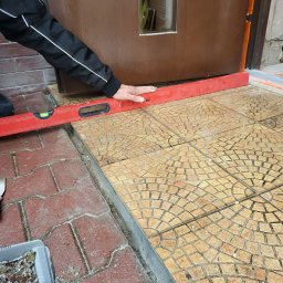 ARBUD Artur Buza - Układanie płytek na zewnątrz budynku przy drzwiach wejściowych. Czerwona poziomica w dłoniach sprawdza równość powierzchni. Widoczne płytki o wzorze mozaiki.