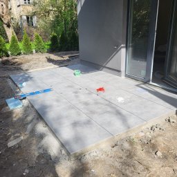 ARBUD Artur Buza - Układanie szarych płytek na tarasie przy domu. Widoczny poziomica, pojemniki z akcesoriami do montażu. W tle budynek z oknami i zieleń.