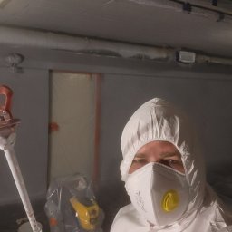 ARBUD Artur Buza - Pracownik w kombinezonie ochronnym i masce trzyma pistolet natryskowy w pomieszczeniu przygotowanym do malowania, z folią ochronną na ścianach i suficie.