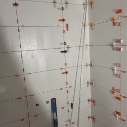 ARBUD Artur Buza - Białe płytki z wzorem w jodełkę, poziomica i system poziomowania płytek w narożniku. Widoczne detale montażu i układania płytek, podkreślające precyzję wykonania.