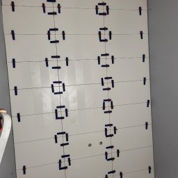 ARBUD Artur Buza - Ściana z białych, połyskliwych płytek, z widocznymi systemami poziomowania płytek, ułożonych w rzędach, w trakcie prac wykończeniowych w rogu pomieszczenia.