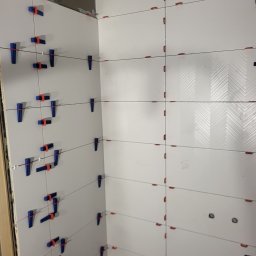 ARBUD Artur Buza - Białe płytki z wzorem w jodełkę, świeżo ułożone na ścianie, z widocznymi niebieskimi i czerwonymi klipsami poziomującymi. W tle fragment podłogi.