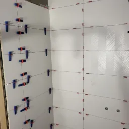 Białe płytki z wzorem w jodełkę, świeżo ułożone na ścianie, z widocznymi niebieskimi i czerwonymi klipsami poziomującymi. W tle fragment podłogi.