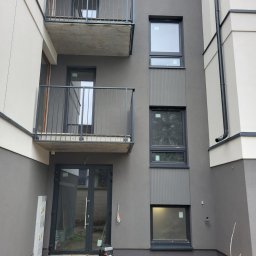 ARBUD Artur Buza - Nowoczesna elewacja budynku wielorodzinnego w odcieniach szarości i bieli z balkonami i oknami, widoczne elementy instalacji zewnętrznej i teren wokół budynku w trakcie prac.