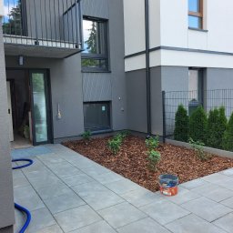 ARBUD Artur Buza - Elewacja budynku po remoncie: szara z białymi akcentami, nowoczesny balkon, okna, wyłożony kostką chodnik, rabata z korą i roślinami. Wiadro z farbą Dulux.