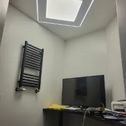 Minimalistyczne wnętrze z grzejnikiem drabinkowym, biurkiem z monitorem i unikalnym oświetleniem sufitowym w kształcie kwadratu z obwódką LED.