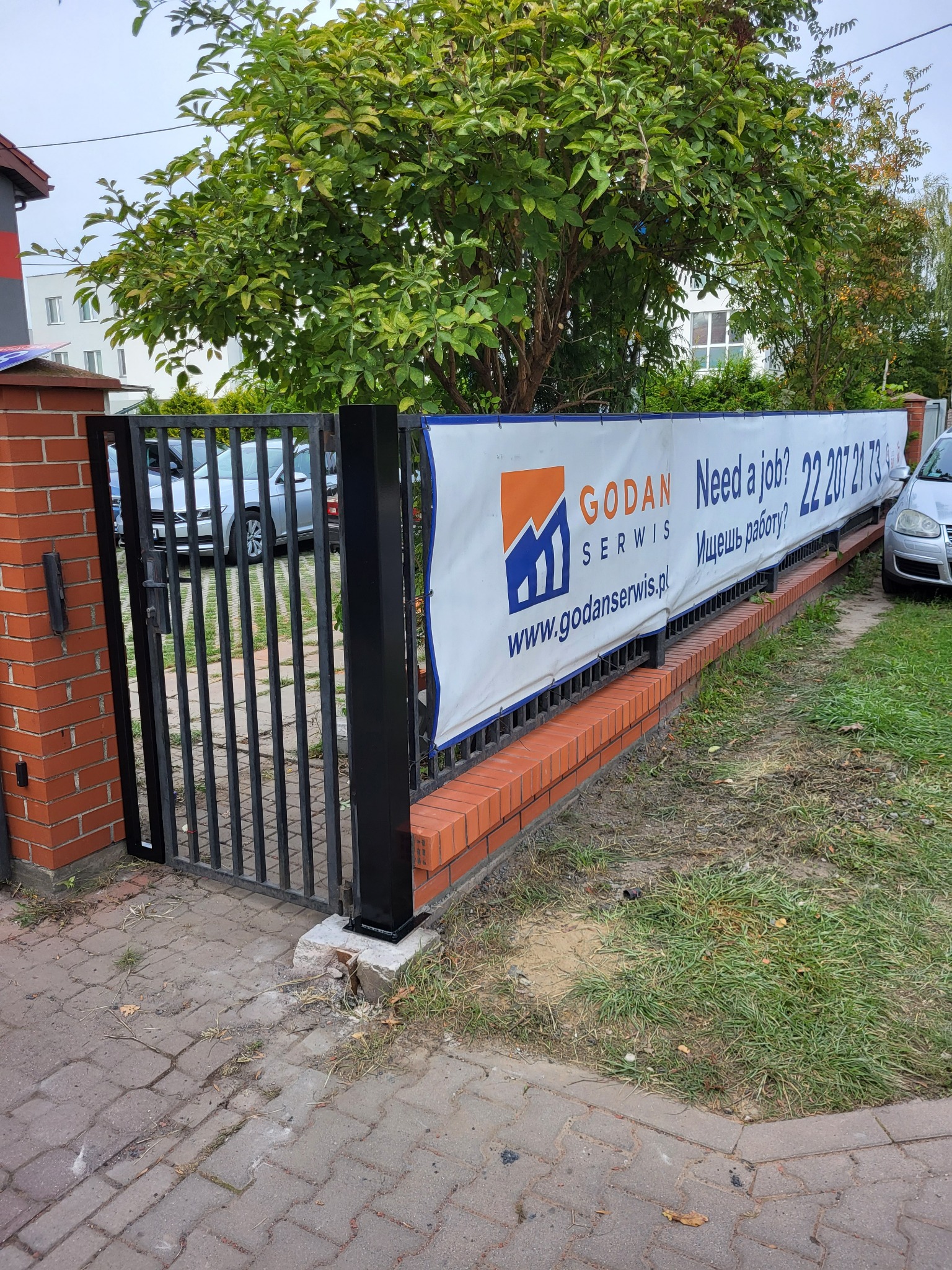 Ogrodzenie z cegły i metalu z banerem reklamowym firmy Godan Serwis, oferującej pracę. Widoczne auta i zieleń w tle. Ujęcie z poziomu chodnika.
