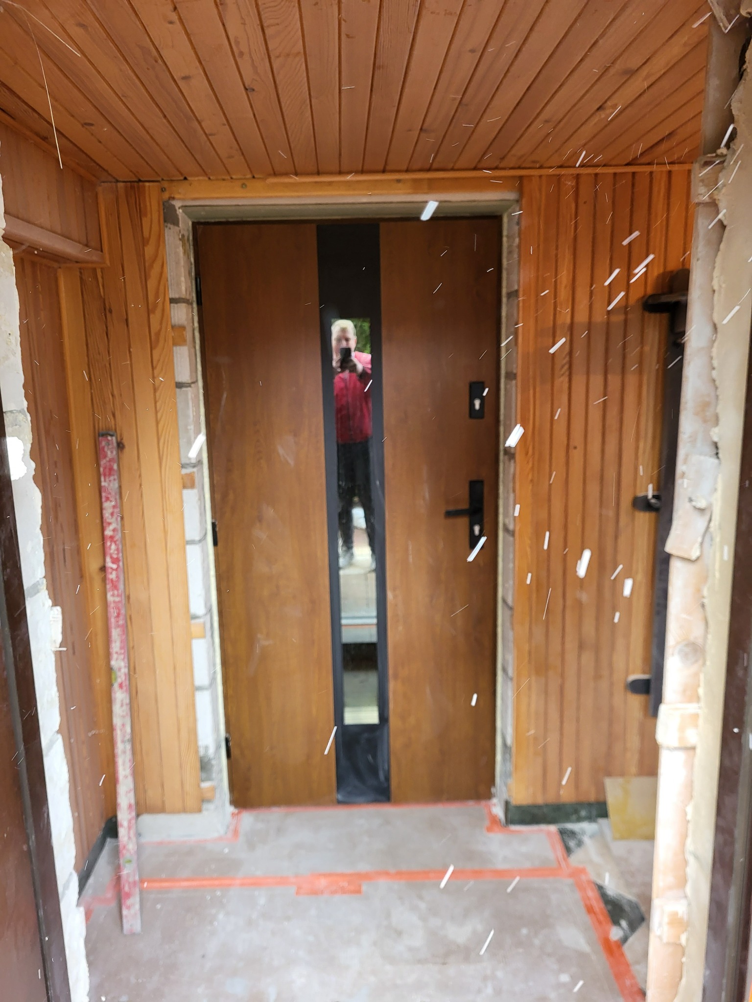 Montaż brązowych drzwi wejściowych z czarnym, pionowym przeszkleniem. Widoczne drewniane panele ścienne i poziomica. Osoba fotografująca odbija się w szybie.