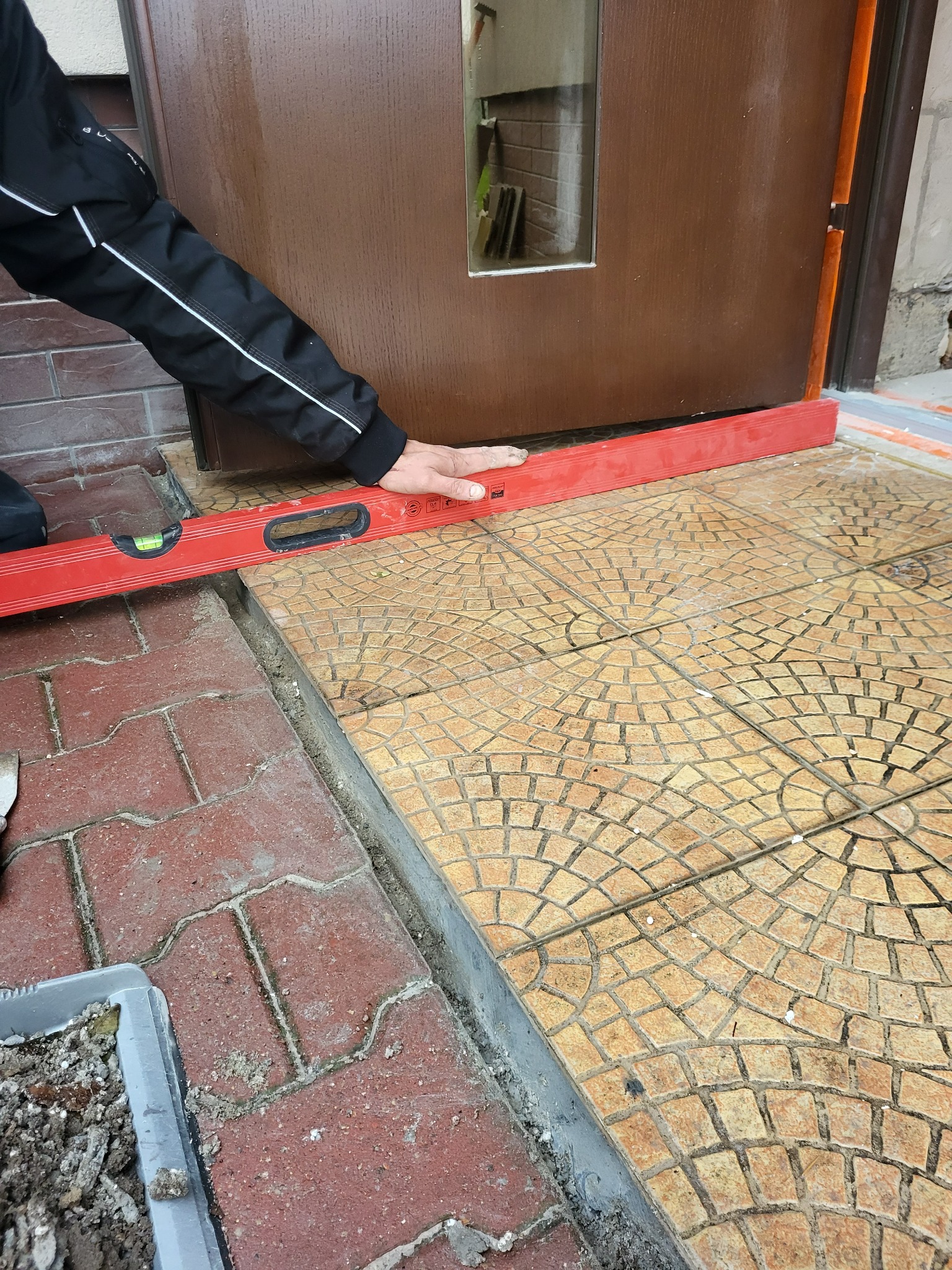 Układanie płytek na zewnątrz budynku przy drzwiach wejściowych. Czerwona poziomica w dłoniach sprawdza równość powierzchni. Widoczne płytki o wzorze mozaiki.