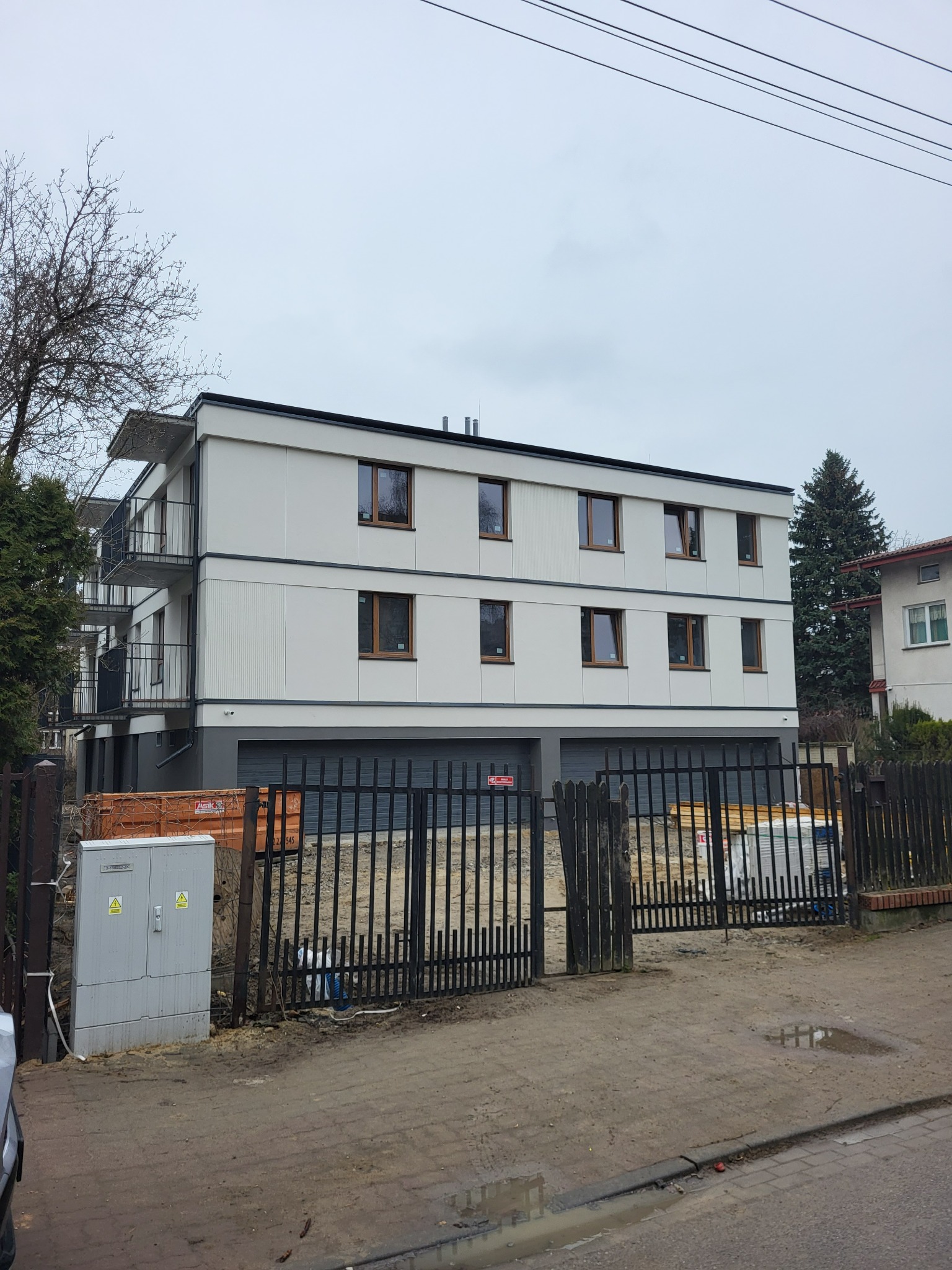 Nowoczesny, trzypiętrowy budynek mieszkalny z balkonami, garażami i ogrodzeniem. Widoczne okna z brązowymi ramami i elementy wykończeniowe.