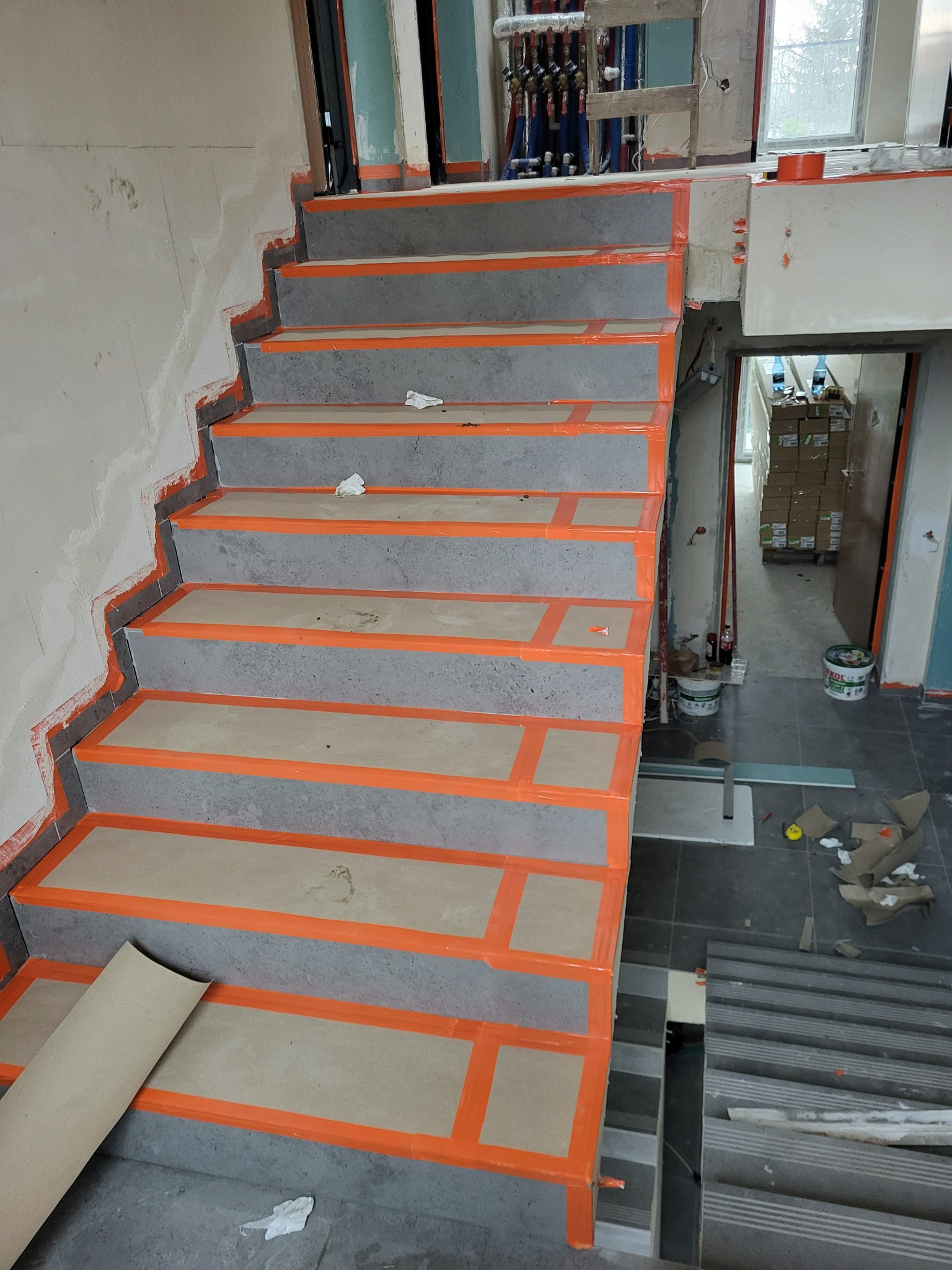Betonowe schody w trakcie wykańczania, zabezpieczone pomarańczową taśmą. Widoczne rury instalacji i materiały budowlane. Etap prac wykończeniowych w surowym wnętrzu.