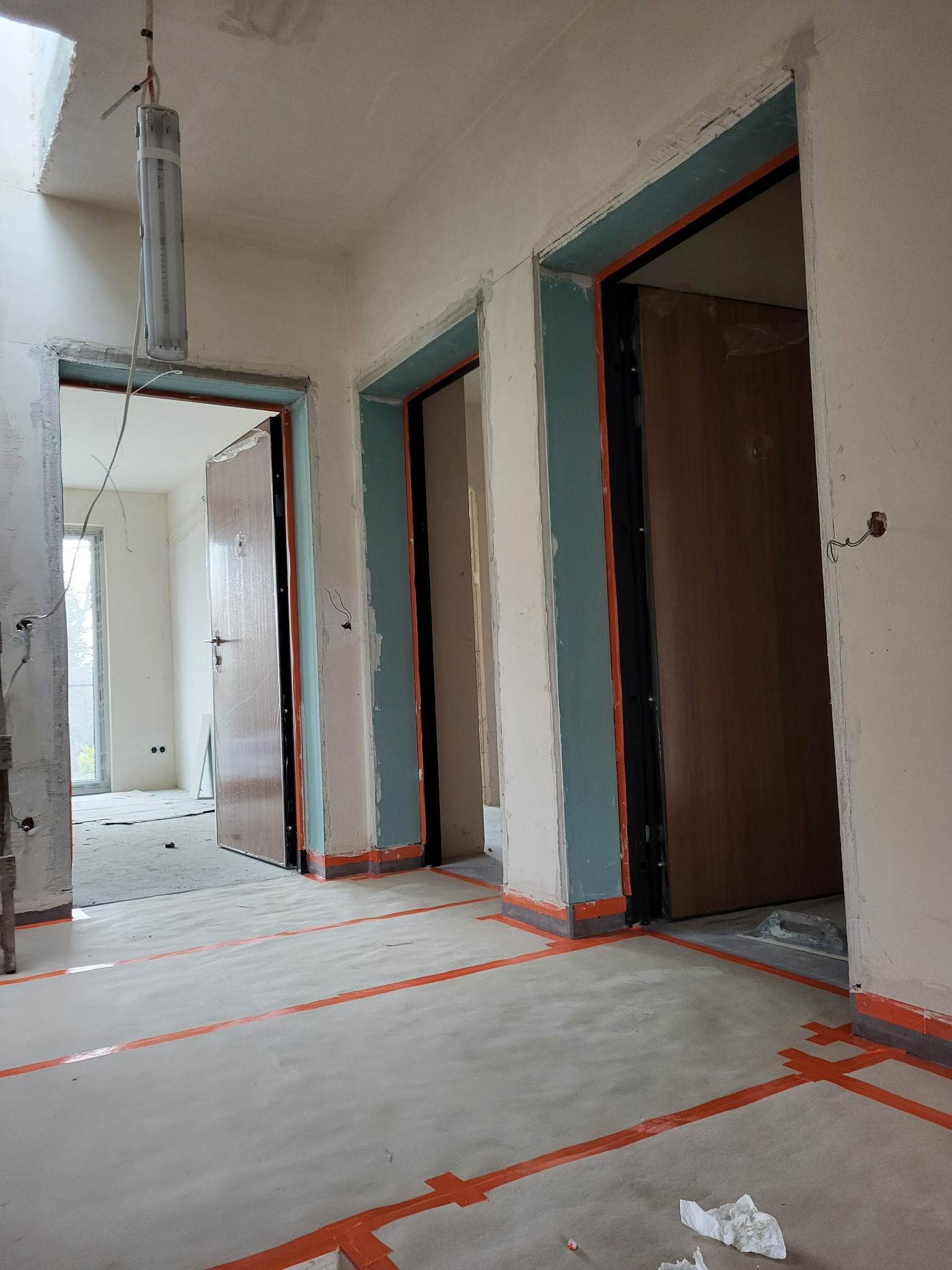 Wnętrze w trakcie prac wykończeniowych: surowe ściany, zabezpieczona podłoga taśmą, widoczne ościeżnice drzwiowe w kolorze turkusowym. W tle okno.