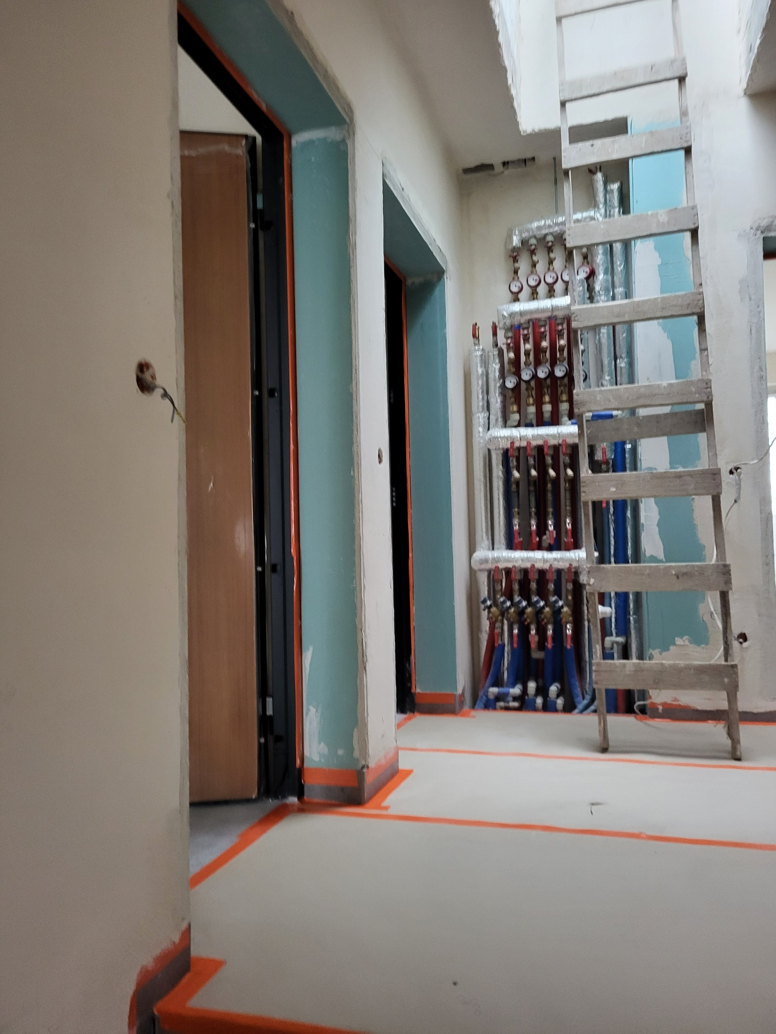 Wnętrze w trakcie wykończenia: widoczne rury instalacji grzewczej, drabina, surowe ściany z otworami na kable i przygotowane ościeżnice drzwiowe z pomalowanymi na niebiesko fragmentami.