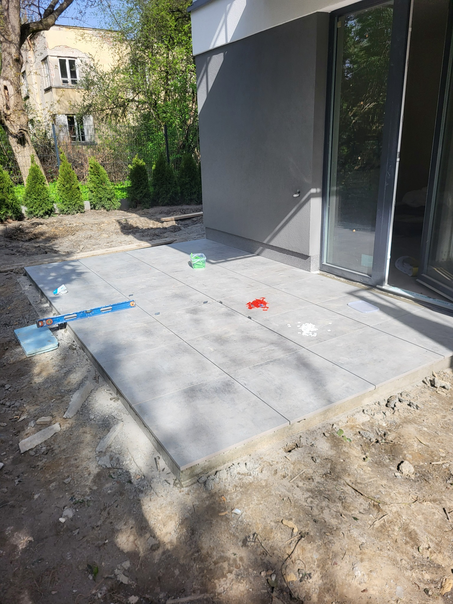 Układanie szarych płytek na tarasie przy domu. Widoczny poziomica, pojemniki z akcesoriami do montażu. W tle budynek z oknami i zieleń.