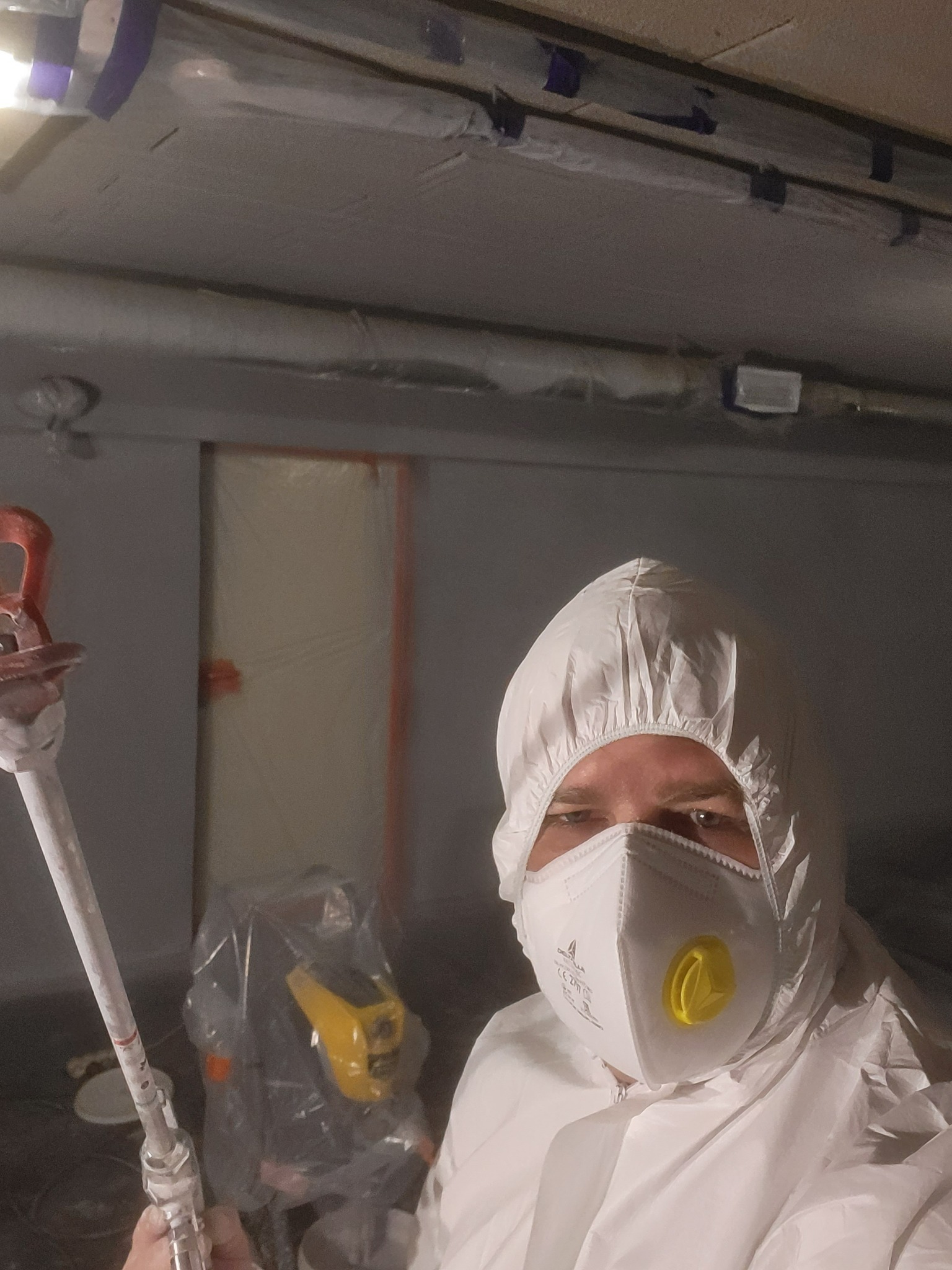 Pracownik w kombinezonie ochronnym i masce trzyma pistolet natryskowy w pomieszczeniu przygotowanym do malowania, z folią ochronną na ścianach i suficie.