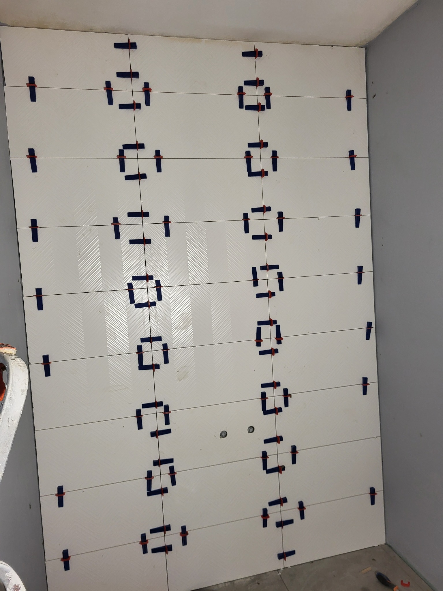 Ściana z białych, połyskliwych płytek, z widocznymi systemami poziomowania płytek, ułożonych w rzędach, w trakcie prac wykończeniowych w rogu pomieszczenia.