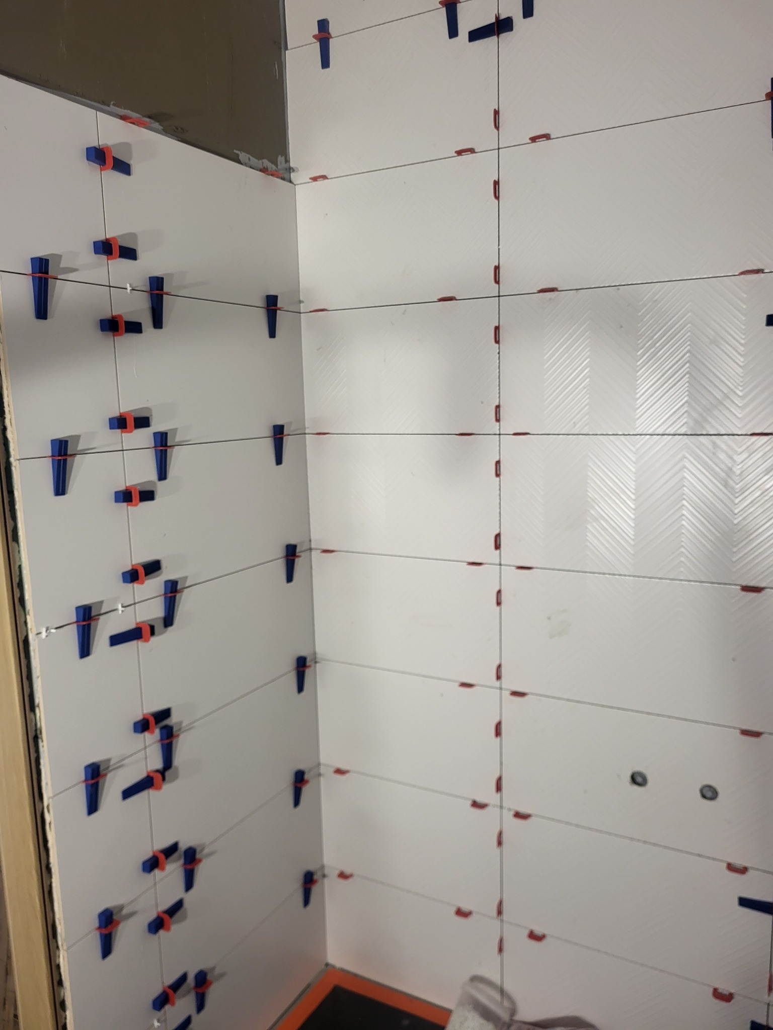 Białe płytki z wzorem w jodełkę, świeżo ułożone na ścianie, z widocznymi niebieskimi i czerwonymi klipsami poziomującymi. W tle fragment podłogi.