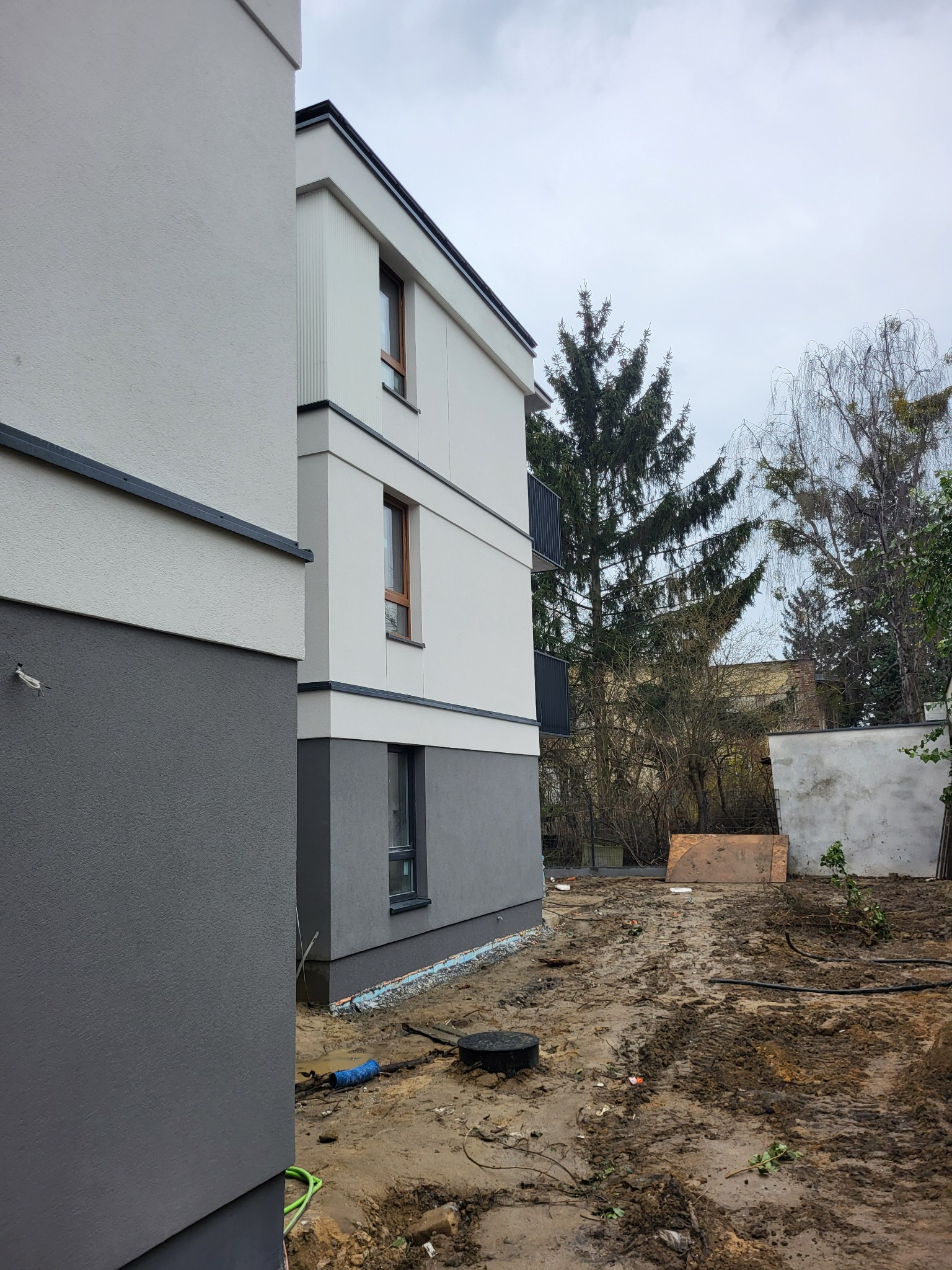 Elewacja budynku wielorodzinnego w Młochowie, z widocznym fragmentem ocieplenia i pomalowaną na szaro dolną częścią. Widoczne okna i fragment terenu.