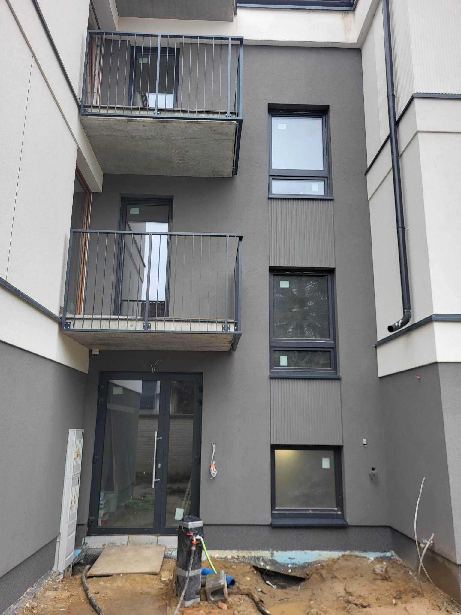 Nowoczesna elewacja budynku wielorodzinnego w odcieniach szarości i bieli z balkonami i oknami, widoczne elementy instalacji zewnętrznej i teren wokół budynku w trakcie prac.