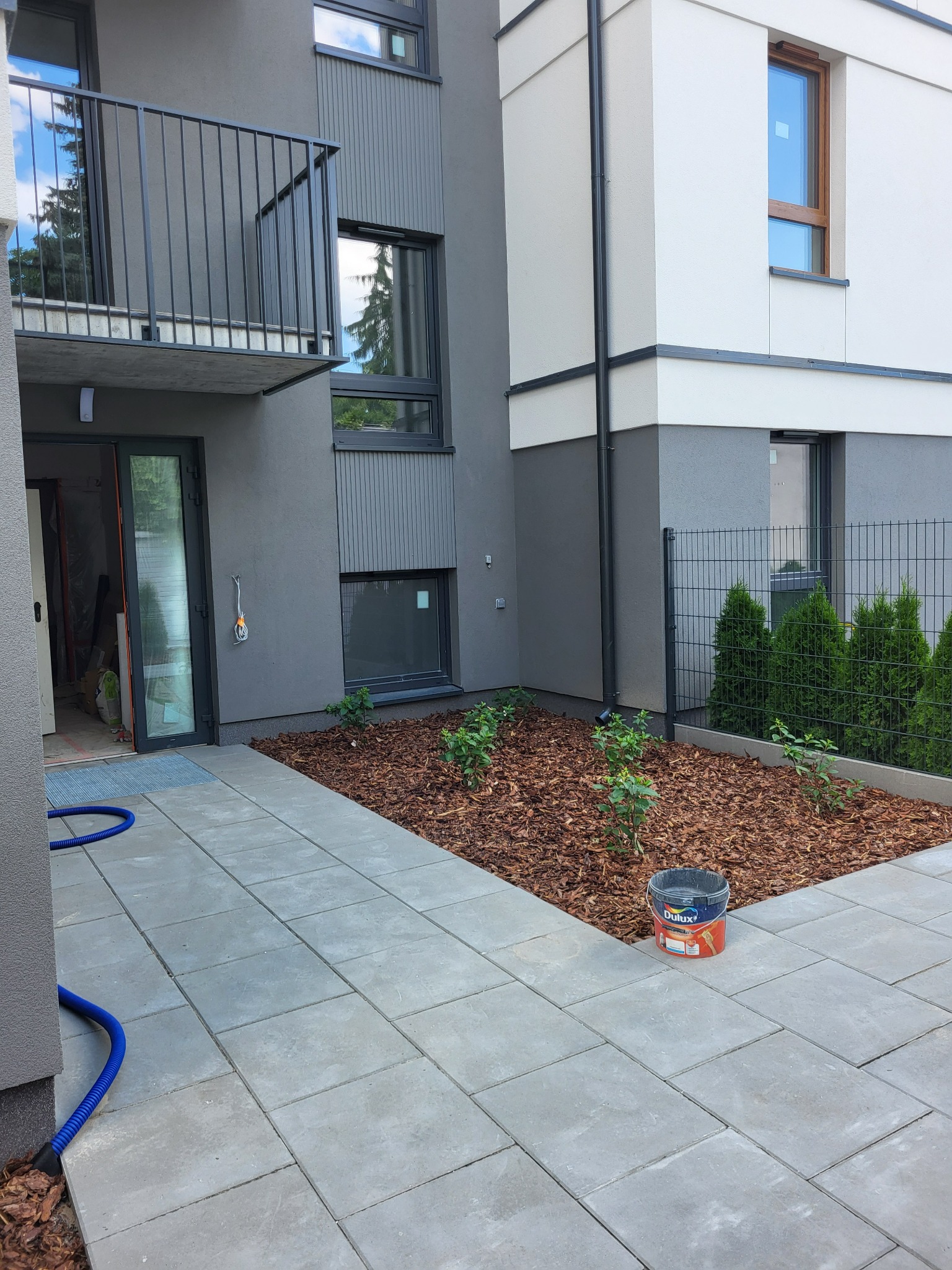 Elewacja budynku po remoncie: szara z białymi akcentami, nowoczesny balkon, okna, wyłożony kostką chodnik, rabata z korą i roślinami. Wiadro z farbą Dulux.
