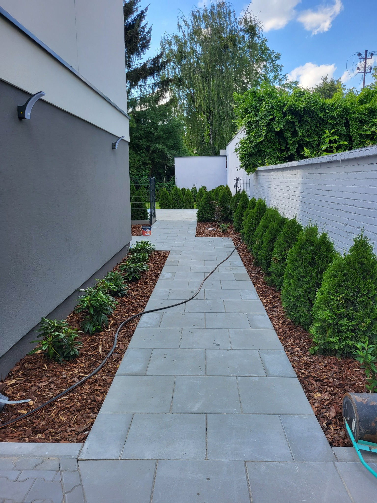 Ścieżka z szarych płyt prowadząca wzdłuż domu i muru, z rabatami wysypanymi korą i obsadzonymi roślinami. Widoczne oświetlenie elewacyjne i fragment nieba.