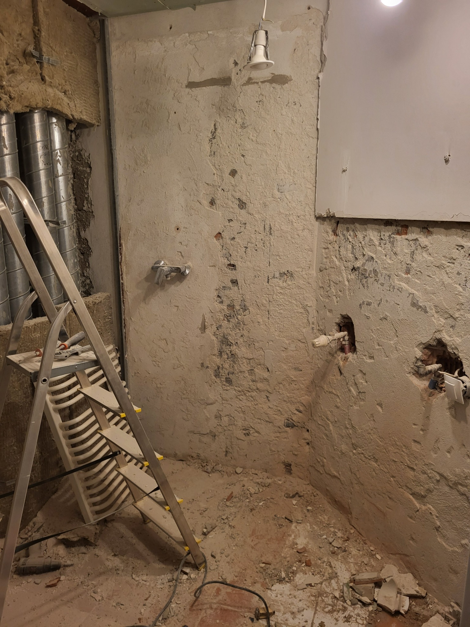 Wnętrze w trakcie remontu: odsłonięte rury, instalacje, surowe ściany z widoczną cegłą, drabina. Widoczne ślady prac remontowych.