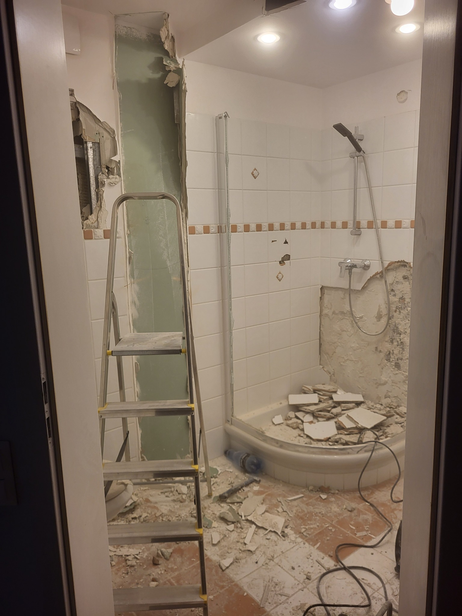 Demontaż płytek w łazience podczas remontu. Widoczna drabina, fragmenty płytek na podłodze, uszkodzone ściany i kabina prysznicowa. Oświetlenie sufitowe.