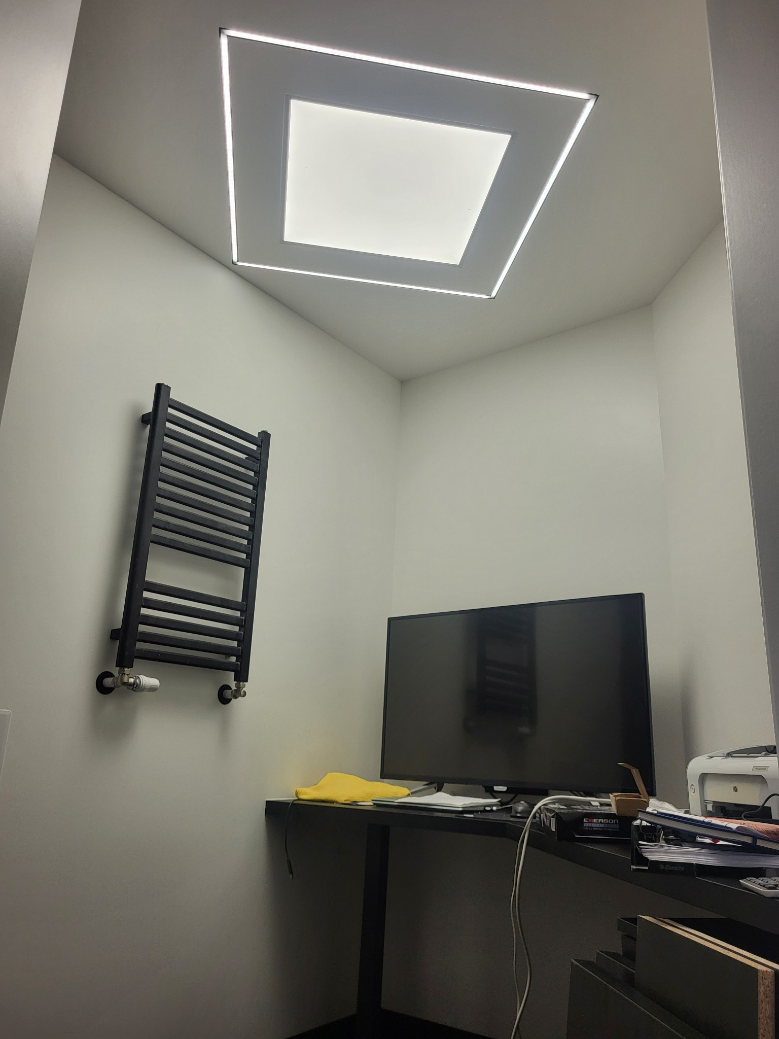 Minimalistyczne wnętrze z grzejnikiem drabinkowym, biurkiem z monitorem i unikalnym oświetleniem sufitowym w kształcie kwadratu z obwódką LED.