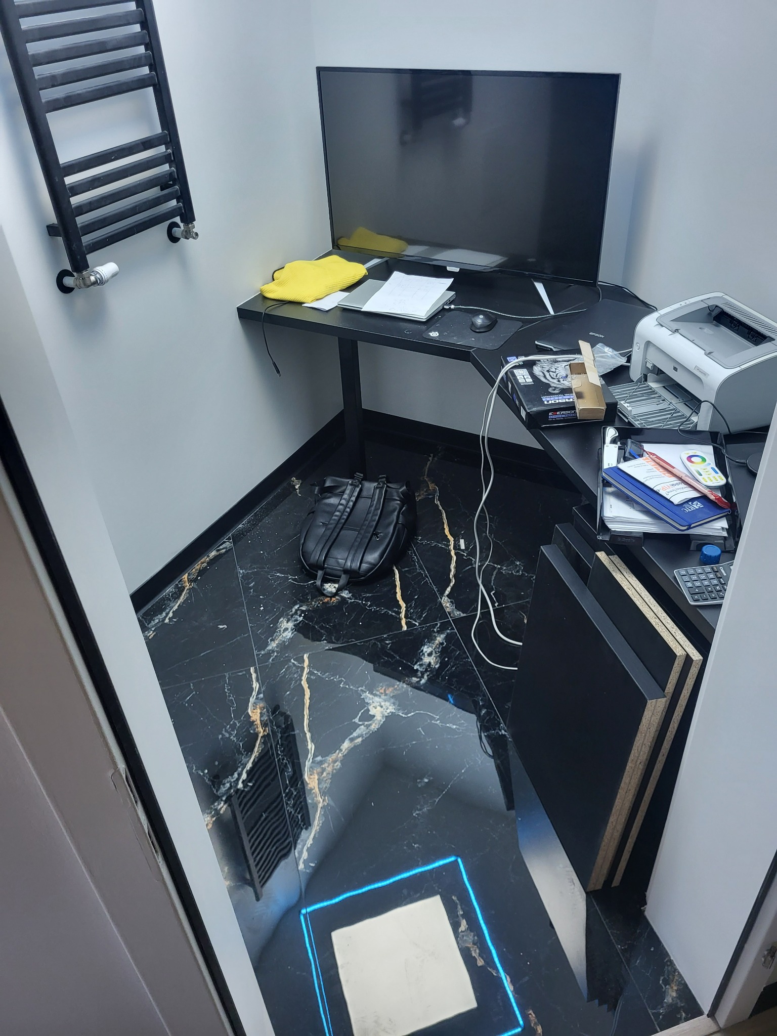 Wnętrze małego pokoju z czarną, marmurkową podłogą i biurkiem narożnym. Na ścianie grzejnik drabinkowy, a na biurku monitor, drukarka i akcesoria biurowe.