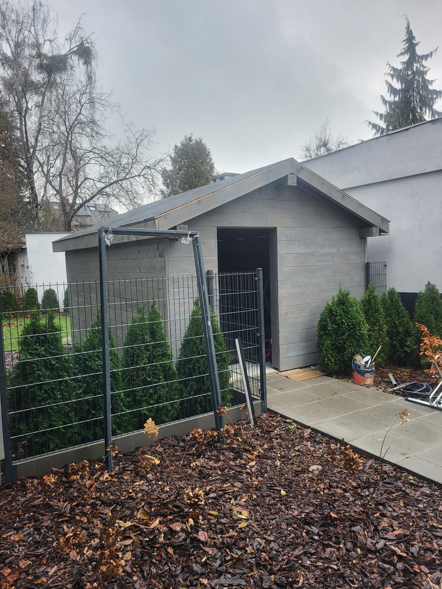 Szary, drewniany domek narzędziowy za czarnym ogrodzeniem panelowym, otoczony tujami i korą. Widoczny kubeł farby i narzędzia malarskie.