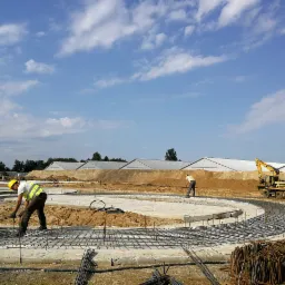 Praca zbrojarza przy tworzeniu okrągłego fundamentu pod budynek, widoczna siatka zbrojeniowa, pracownicy w kaskach i odblaskowych kamizelkach, w tle budynek hali i koparka CAT.