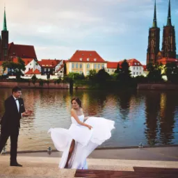 Para młoda podczas sesji zdjęciowej we Wrocławiu na tle Odry i katedry, panna młoda w białej sukni z rozcięciem, pan młody w czarnym smokingu.